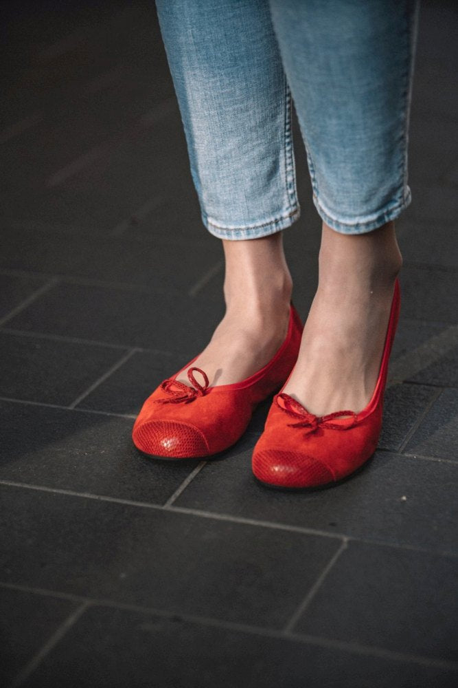 Eine Person trägt ZAQQs TWIST Velours Red Ballerinas mit Schleife und Kautschuksohle, kombiniert mit hellblauen Cropped Jeans, und steht anmutig auf einem dunklen Fliesenboden.