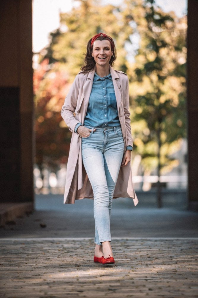 Eine Frau geht selbstbewusst an einem sonnigen Tag im Freien spazieren. Sie trägt einen leichten Trenchcoat, ein Jeanshemd und Jeans. Sie lächelt, hat ein rotes Stirnband im Haar und TWIST Velours Red-Schuhe von ZAQQ schmücken ihre Füße vor einem Hintergrund aus verschwommenen Bäumen.