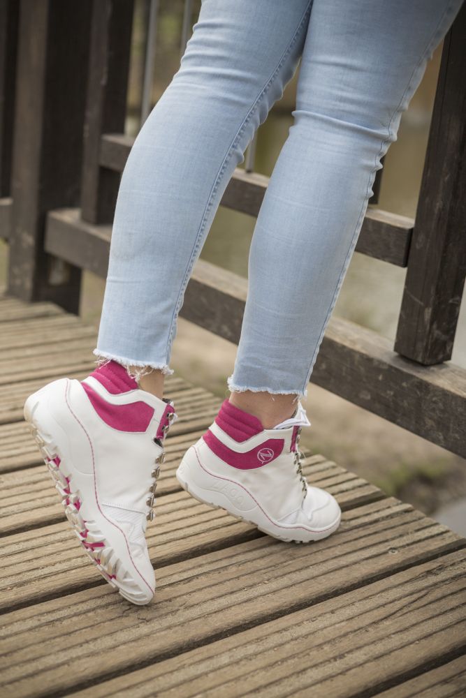 Eine Person in hellblauer Jeans und weißen Sneakers mit pinken Akzenten, möglicherweise den ZAQQ VAQATION Pink Waterproof, steht auf einem Holzdeck neben einem Geländer. Ihre Haltung lässt vermuten, dass sie auf ein Gewässer blickt.