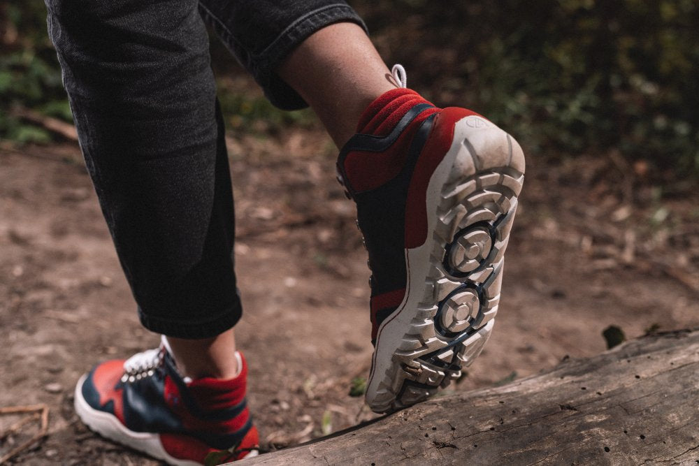 Eine Person in schwarzen Jeans und Sneakers mit ZAQQ VAQATION Red Waterproof-Design steigt auf einem Feldweg über einen Baumstamm. Das Profil der Sneakers ist deutlich sichtbar und unterstreicht ihre Strapazierfähigkeit und Griffigkeit.