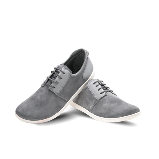 Die Barfuß-Sneaker PIQUANT Grey von ZAQQ aus grauem Wildleder mit flexibler Sohle werden vor einem weißen Hintergrund präsentiert. Ein Schuh steht aufrecht, während der andere geneigt ist, wodurch die Schnürsenkel und die glatte Oberfläche hervorgehoben werden.